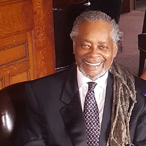 Baba Charles Simmons smiling in a dark suit, patterned tie, and long gray locs.